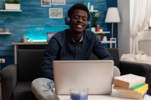 Black student wearing headphone having audio business course on laptop computer using univeristy e-learning platform in living room. Afro teenager smiling while listening online music enjoying leisure